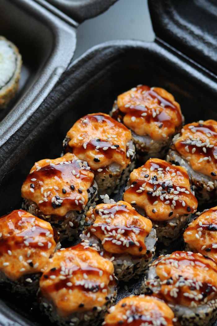 about-img Delicious spicy sushi rolls topped with sesame seeds and sauce in a takeout container.